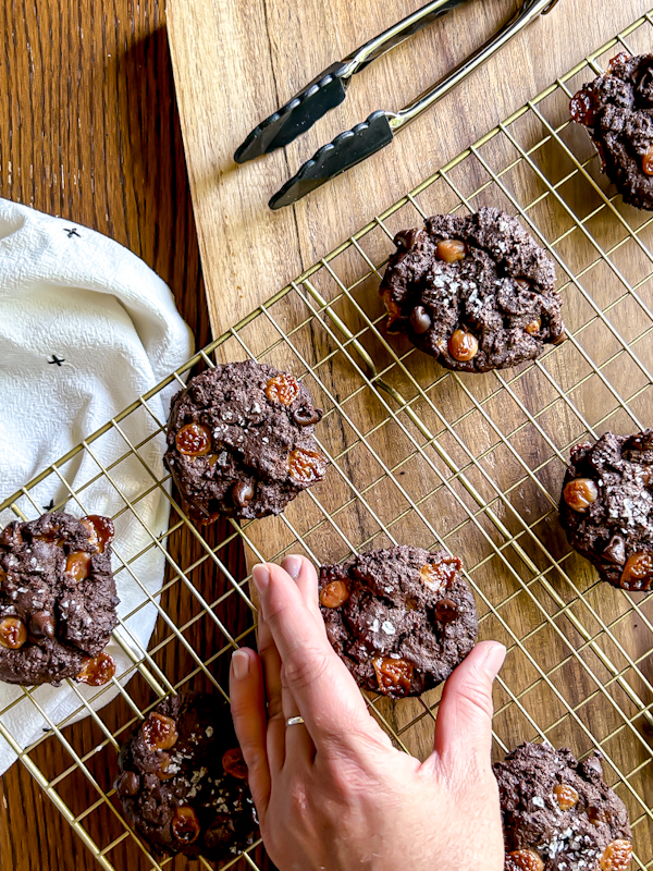 A hand reaches for a double chocolate chip cookie cooling on a gold metal rack on a wood board with natural window light coming from the side of the image.